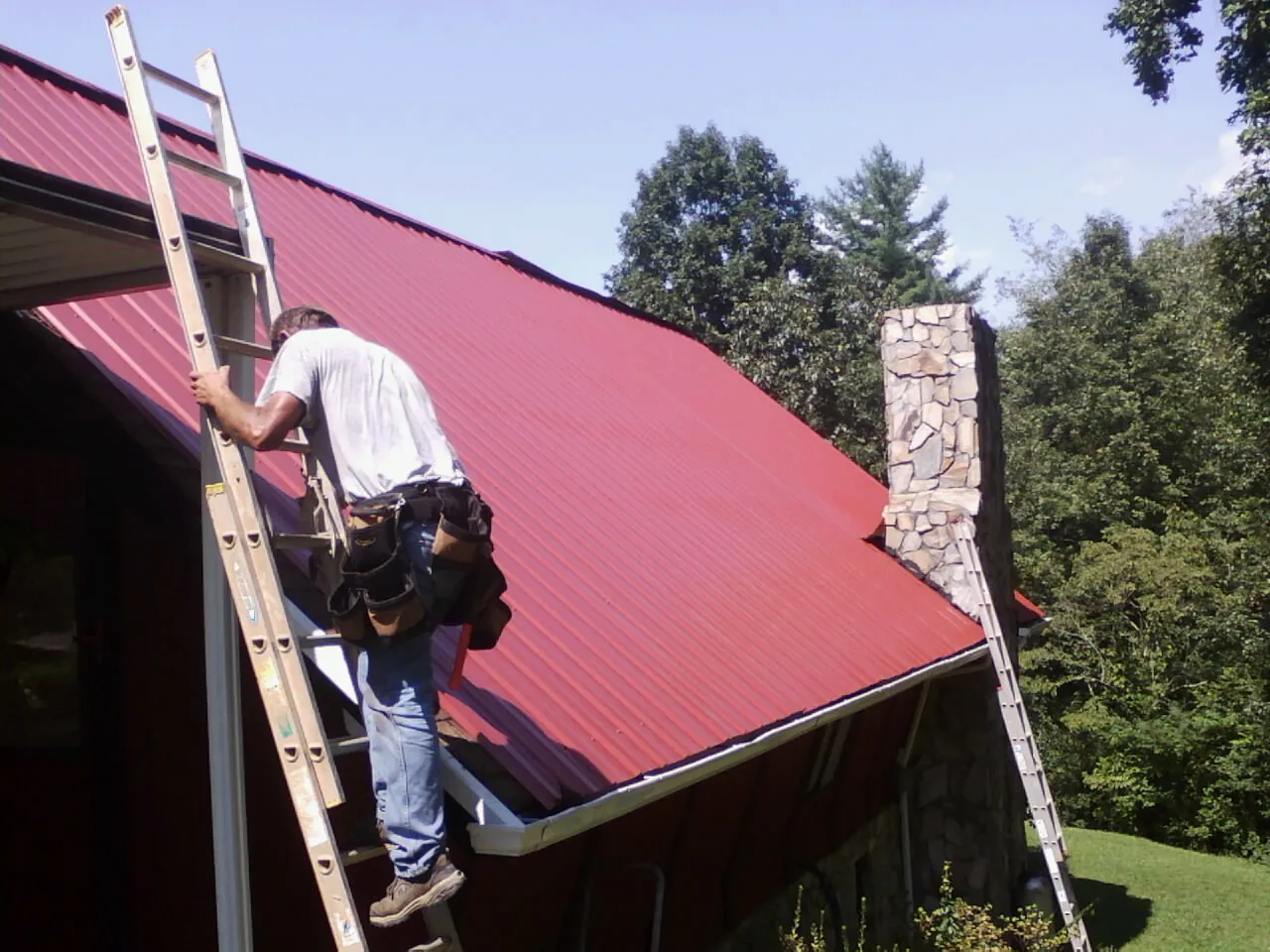 Roofer on a ladder beside a red metal roof and stone chimney — Soffit & Fascia Repair by Roofing Brothers in Geigertown